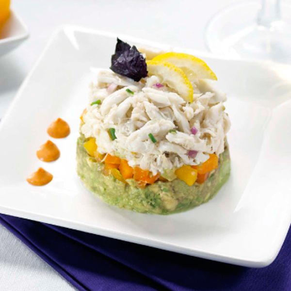 A layered crab salad with diced vegetables and avocado, garnished with lemon slices and a purple leaf on a white plate.