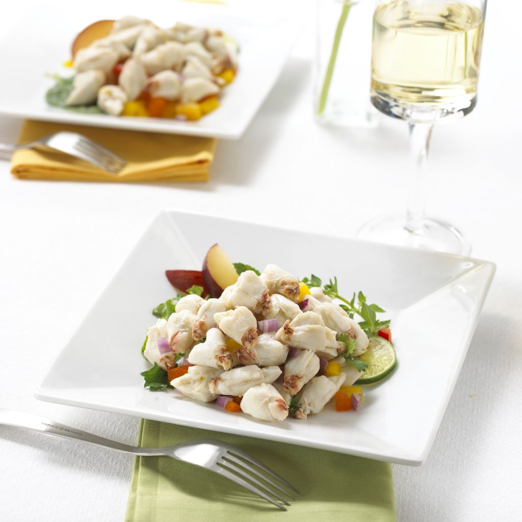 Crab salad with greens and sliced fruit on a square plate, served with white wine on a white tablecloth.