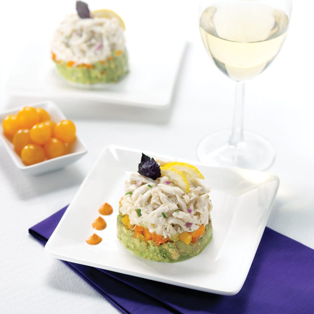 A layered crab salad with avocado and vegetables, garnished with lemon, served beside a glass of white wine.